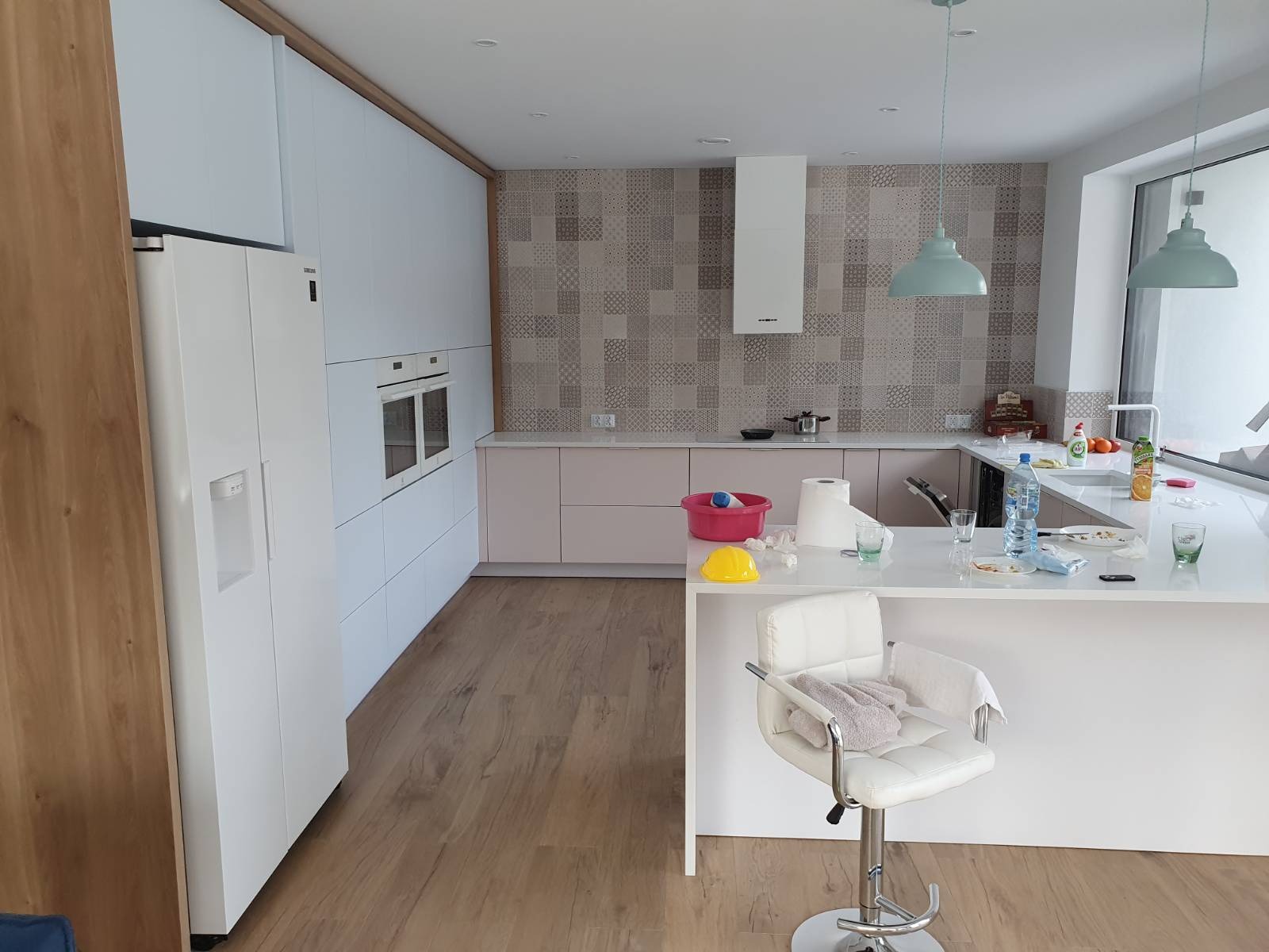 Jasna kuchnia z białymi frontami i drewnianymi akcentami. Widoczny blat z wyspą, krzesło barowe i sprzęt AGD. Na blacie naczynia i akcesoria kuchenne.