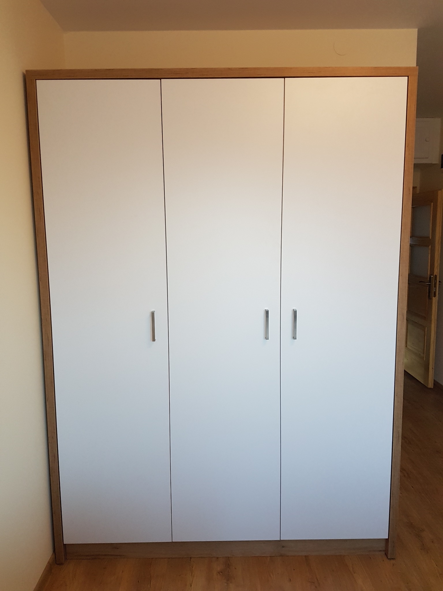 Trzydrzwiowa szafa z białymi frontami i drewnianą ramą, minimalistyczny design, widoczna podłoga z paneli, drzwi do innego pomieszczenia w tle.