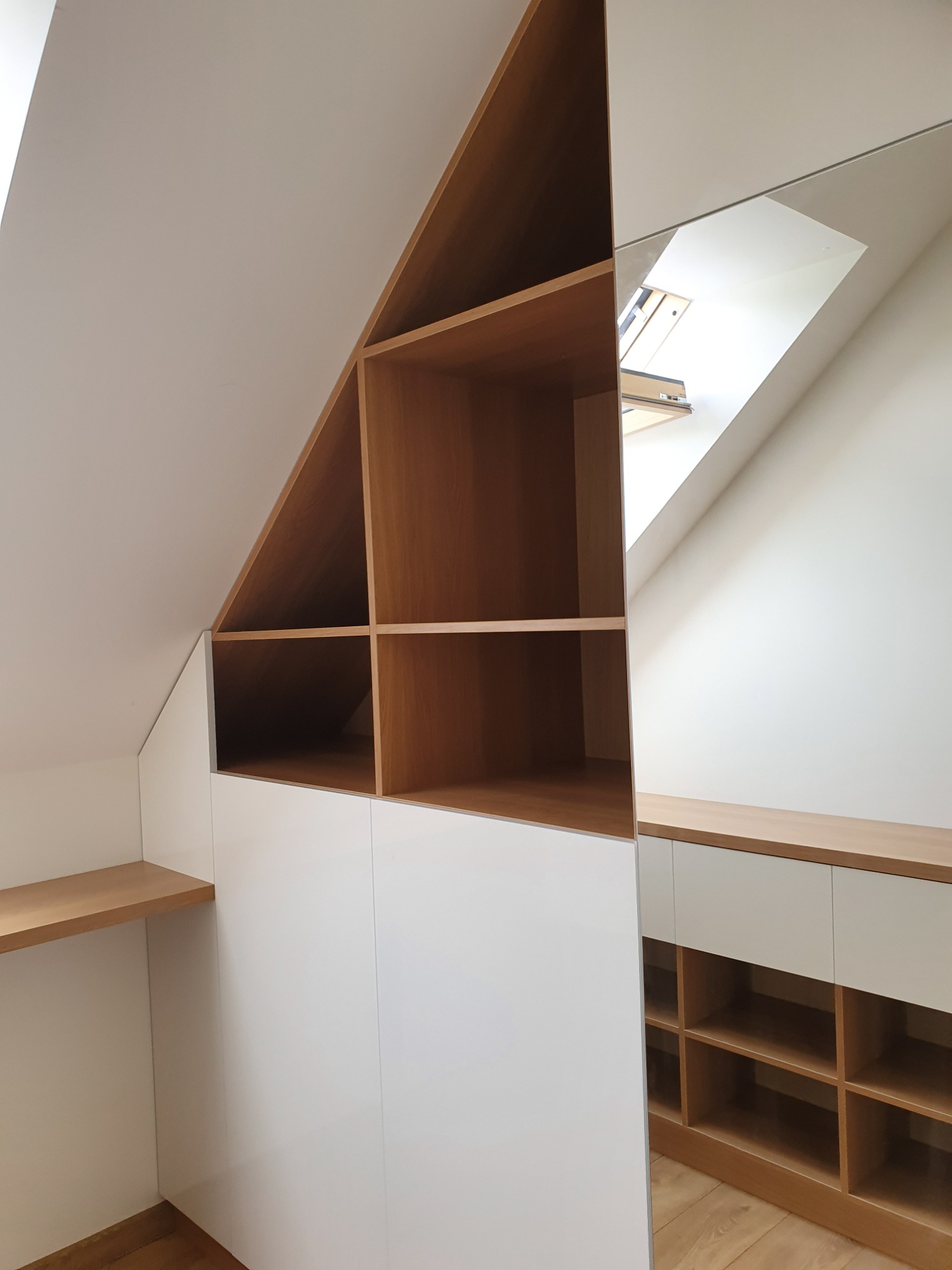 Szafa wnękowa z białymi frontami i drewnianymi półkami, dopasowana do skosu dachu, z lustrzanym odbiciem fragmentu wnętrza i okna dachowego.