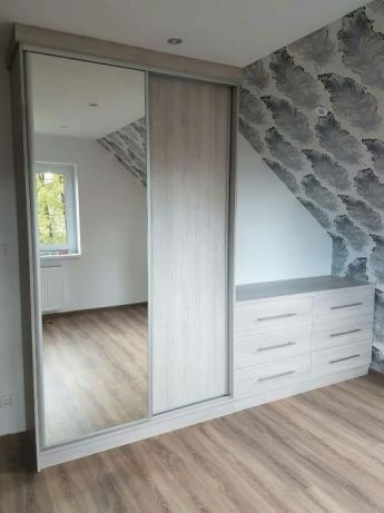 Jasna szafa wnękowa z przesuwnymi drzwiami lustrzanymi, wkomponowana w skos pomieszczenia, z komodą z szufladami po prawej stronie, na tle ściany w pióra.