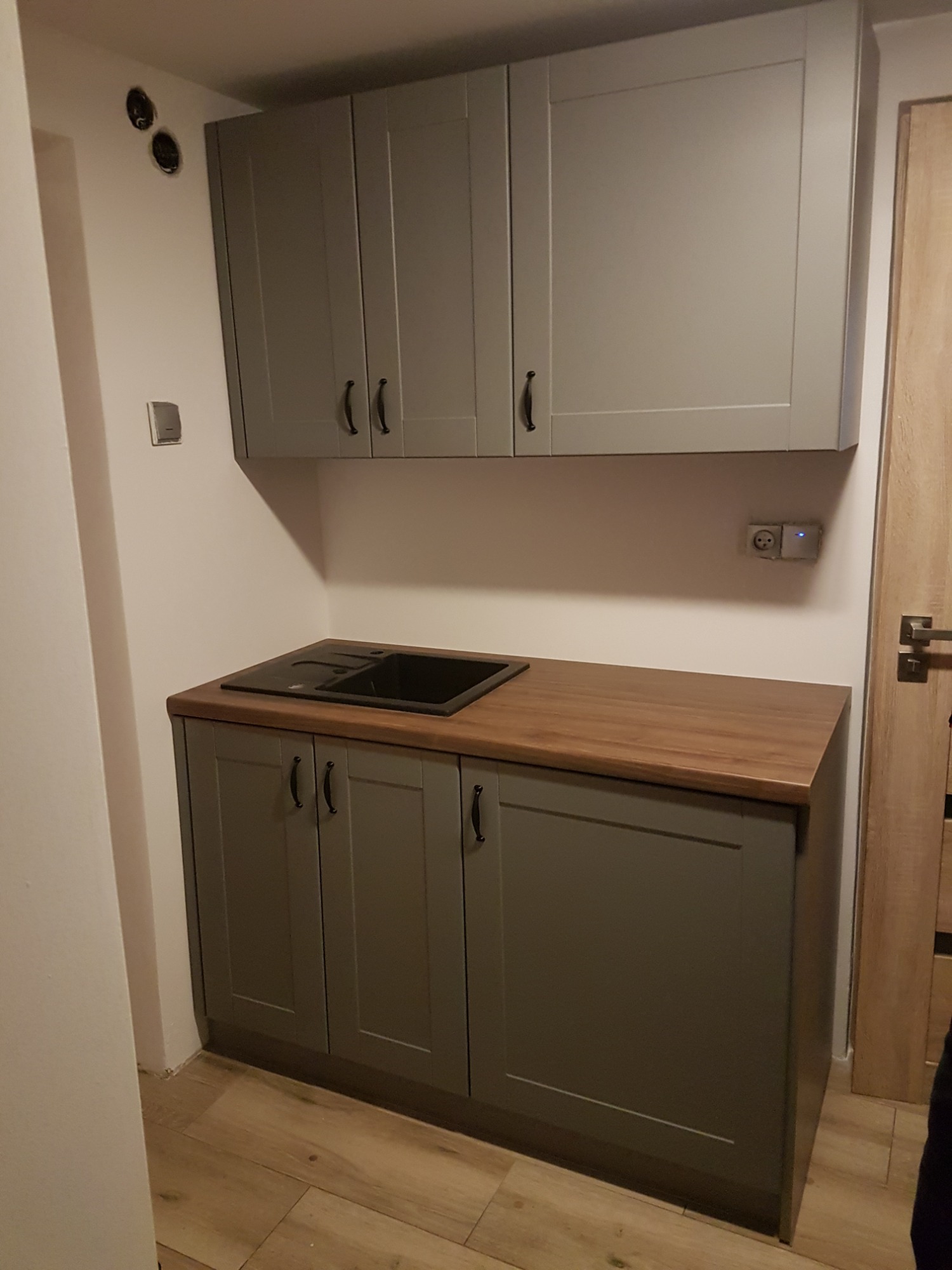 Szara zabudowa kuchenna z drewnianym blatem i czarnym zlewozmywakiem, widoczne gniazdko elektryczne z podświetleniem LED.