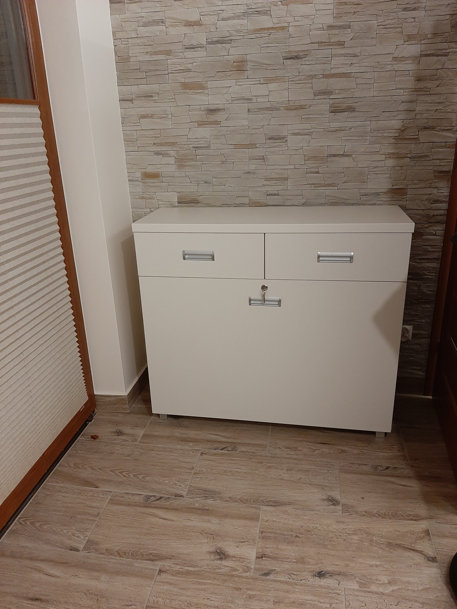Biała komoda z dwoma szufladami i zamykaną szafką na tle ściany wyłożonej kamieniem dekoracyjnym, widoczna na drewnopodobnej podłodze.