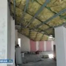 Poddasze w trakcie izolacji termicznej wełną mineralną, widoczne metalowe profile konstrukcyjne, fragment ściany z płyt kartonowo-gipsowych w różowe pasy, surowy stan wykończenia.