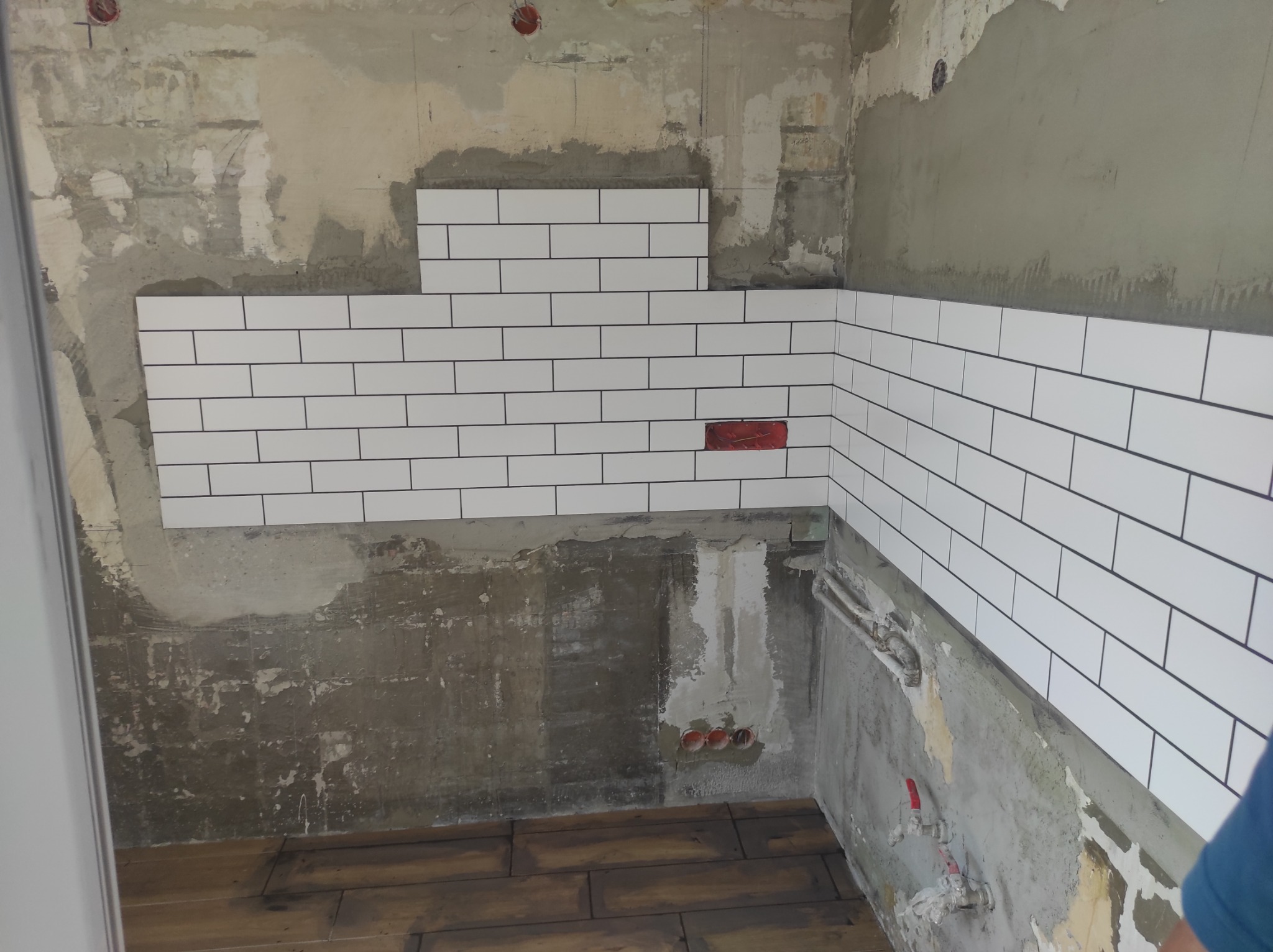 Fragment ściany w trakcie układania białych, podłużnych płytek z czarną fugą, z widocznym surowym tłem ściany i podłogi z paneli, jeden czerwony punkt instalacyjny na ścianie.