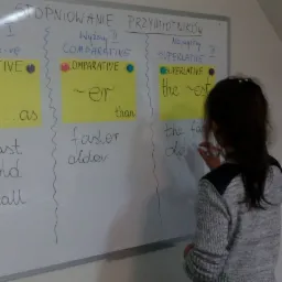 Dziewczynka pisze markerem na białej tablicy z napisem 'Stopniowanie Przymiotników' i przykładami stopniowania przymiotników w języku angielskim.