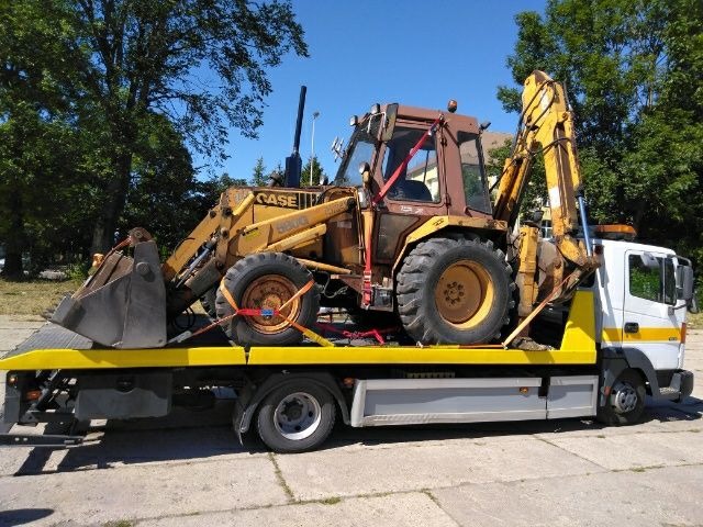 Żółto-brązowa koparko-ładowarka Case 580G zabezpieczona czerwonymi pasami transportowymi na żółtej platformie białej lawety. Widok z boku na tle drzew i błękitnego nieba.