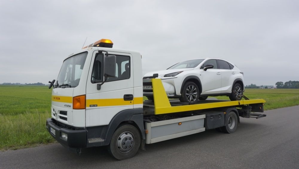 Biała laweta z pomarańczowym światłem ostrzegawczym przewozi białego Lexusa NX na tle zielonego pola i pochmurnego nieba.