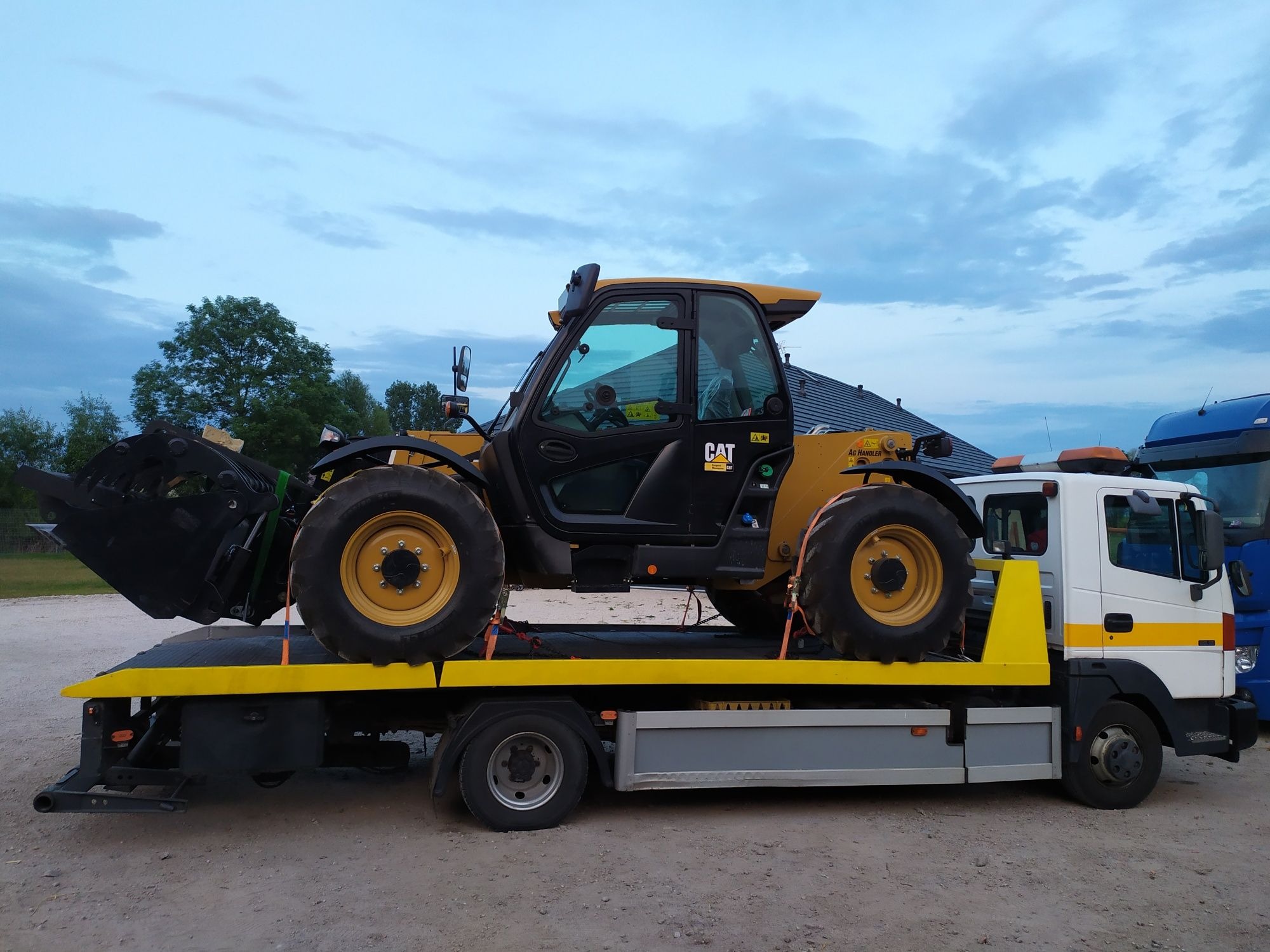 Żółty teleskopowy ładowacz CAT zabezpieczony pasami transportowymi na żółtej platformie lawety. Widoczna kabina operatora i duża łyżka załadowcza. Tło stanowią budynki i drzewa pod pochmurnym niebem.