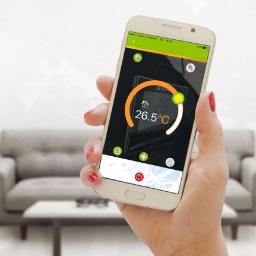 KAMEL Mariusz Kamela - Kobieta trzyma smartfon z aplikacją do zdalnego sterowania temperaturą w pomieszczeniu, wyświetlającą 26.5 stopni Celsjusza. W tle widoczny salon z szarą sofą.