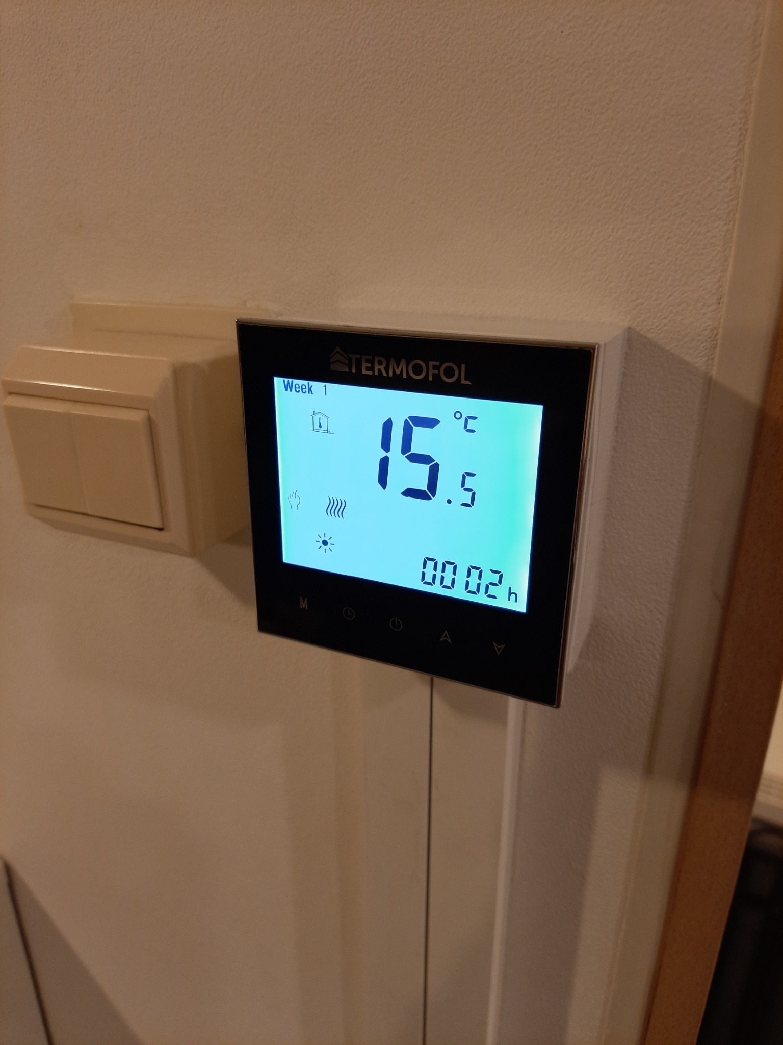 Czarny panel sterowania ogrzewaniem podłogowym marki Termofol zamontowany na beżowej ścianie obok białego włącznika światła, wyświetlający temperaturę 15.5 stopni Celsjusza.