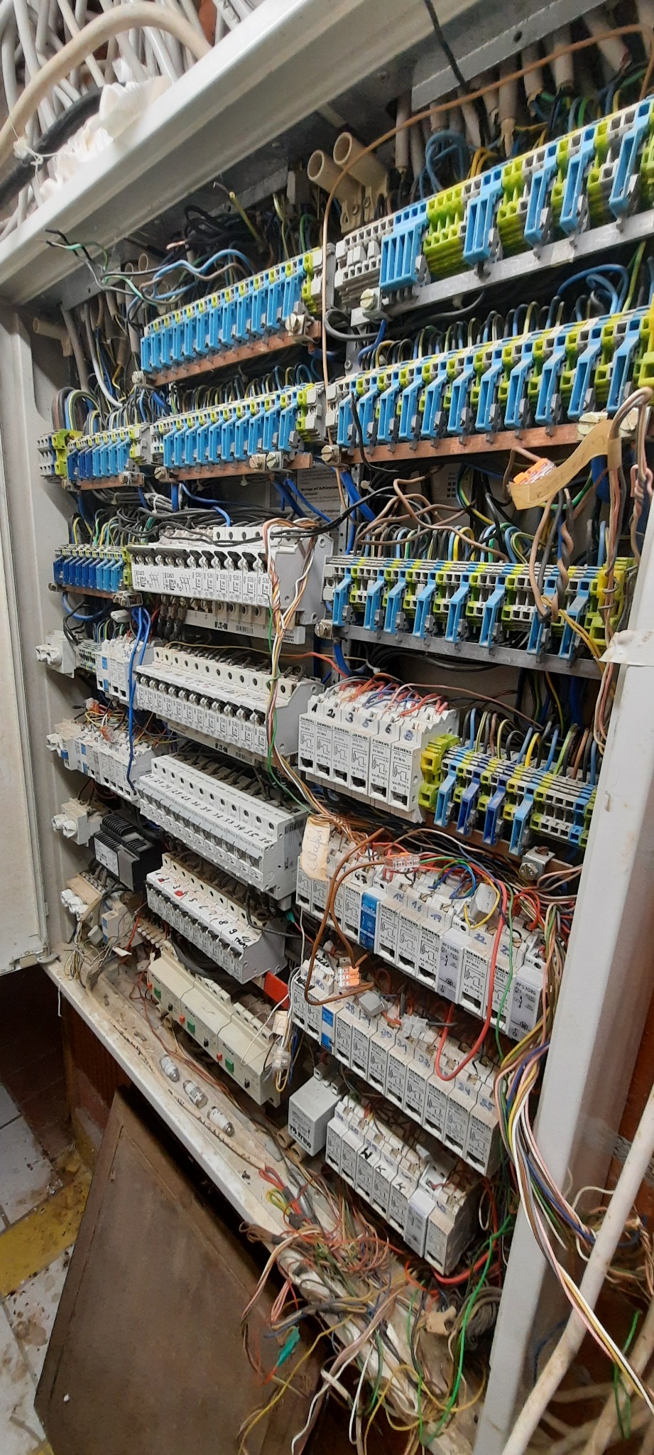 Otwarta skrzynka elektryczna z licznymi przewodami, bezpiecznikami i listwami zaciskowymi, widoczne zabrudzenia na obudowie i podzespołach.