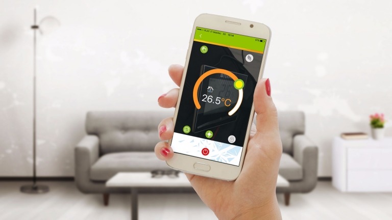 Kobieta trzyma smartfon z aplikacją do zdalnego sterowania temperaturą w pomieszczeniu, wyświetlającą 26.5 stopni Celsjusza. W tle widoczny salon z szarą sofą.