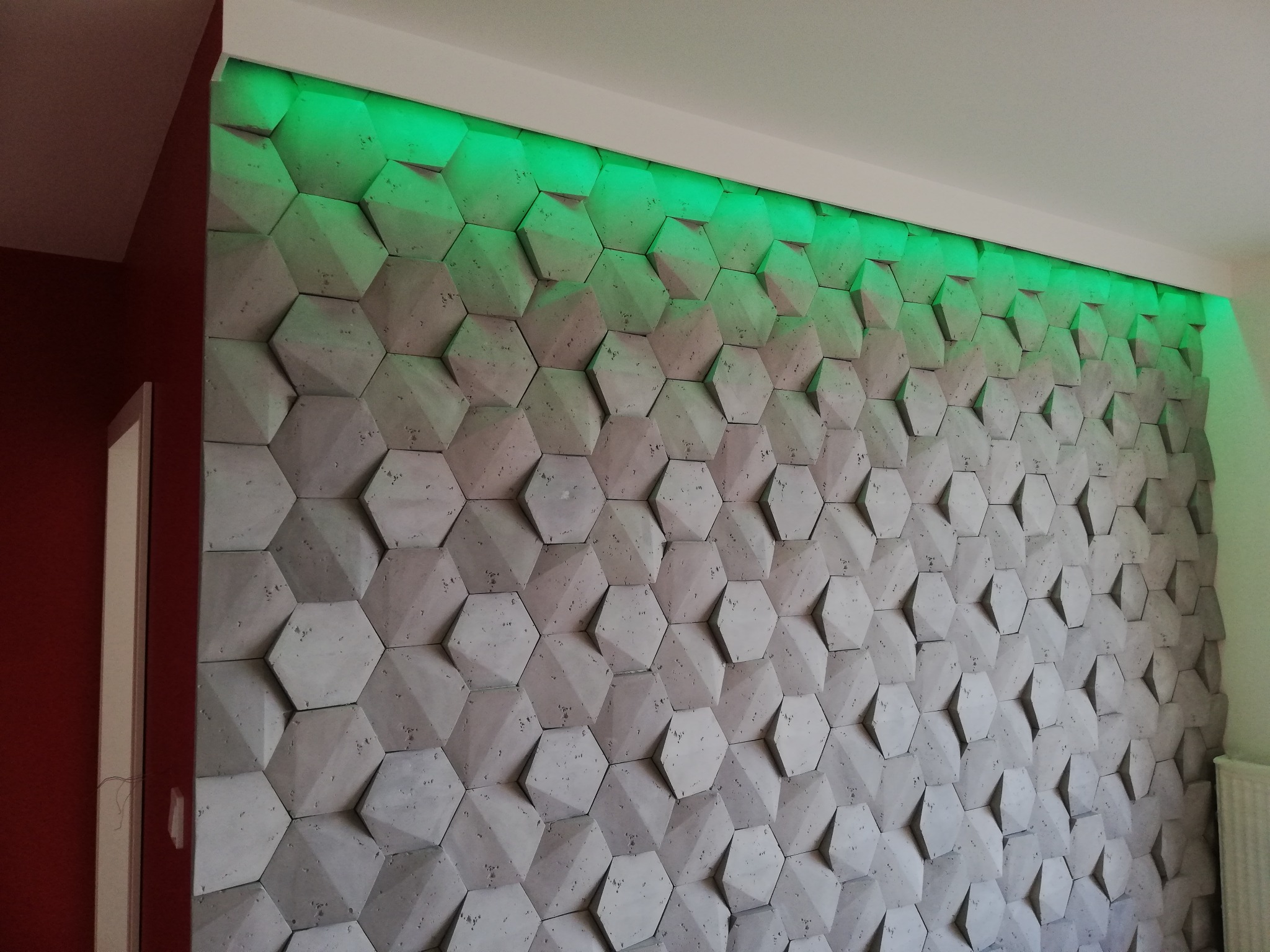Dekoracyjna ściana z betonowych heksagonów oświetlona od góry zielonym światłem LED, widok na część ściany i narożnik pokoju.