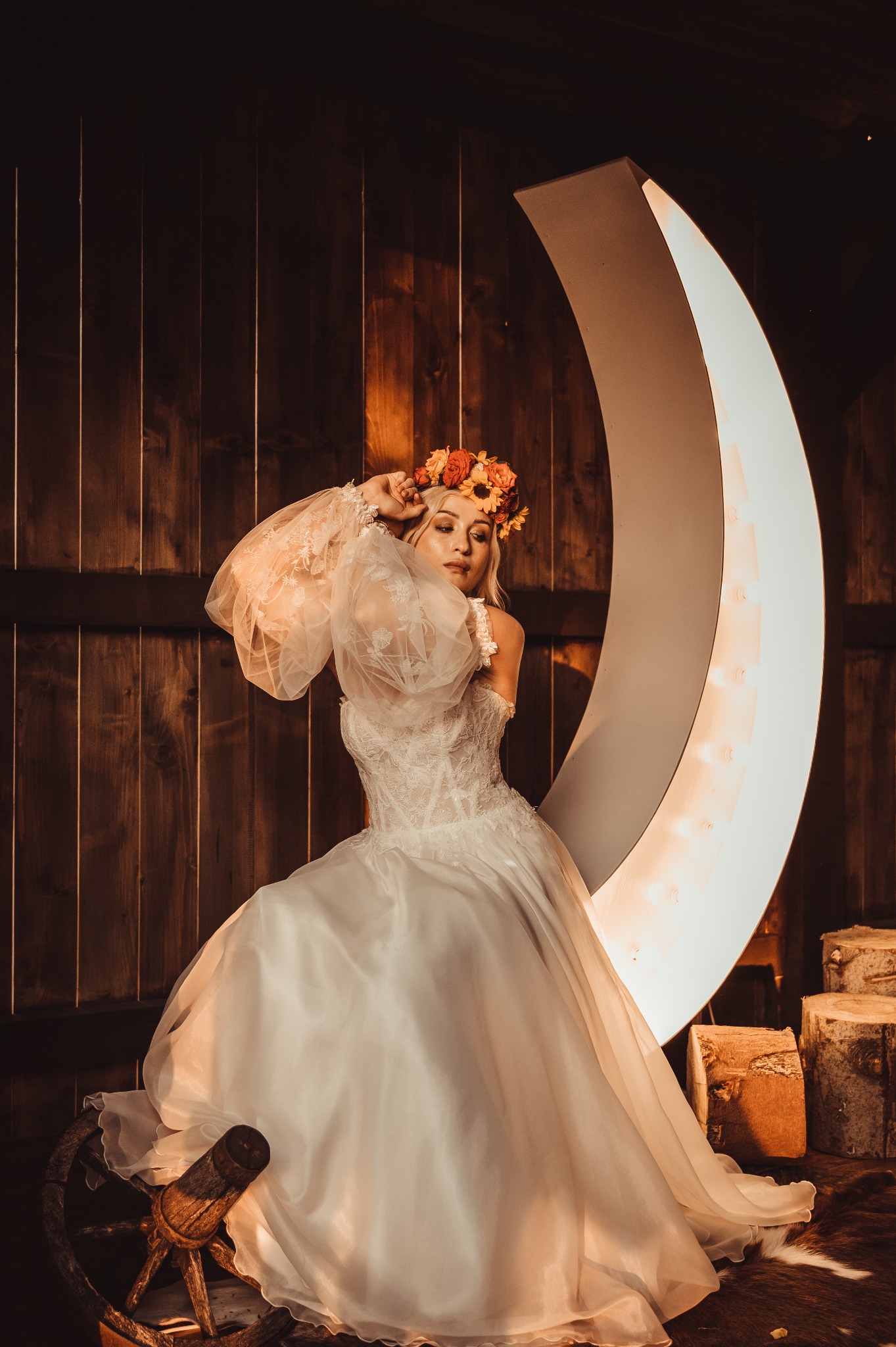 Blondwłosa kobieta w koronkowej sukni ślubnej z bufiastymi rękawami i wiankiem z kwiatów pozuje na tle drewnianej ściany i świecącego księżyca, siedząc na starym wozie.