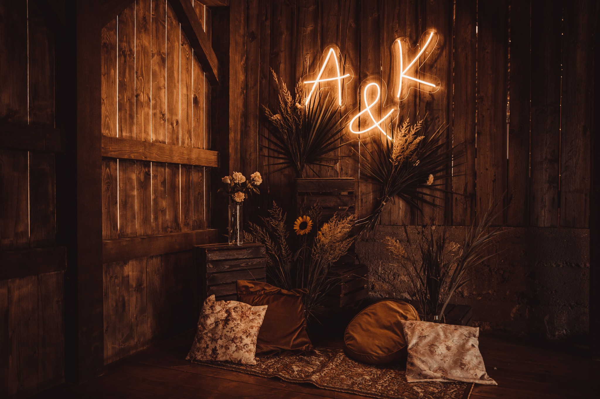 Rustykalna aranżacja ślubna z drewnianym tłem, neonem z inicjałami A&K, kwiatami w wazonie, dekoracyjnymi poduszkami i dywanem w ciepłych odcieniach brązu i beżu.