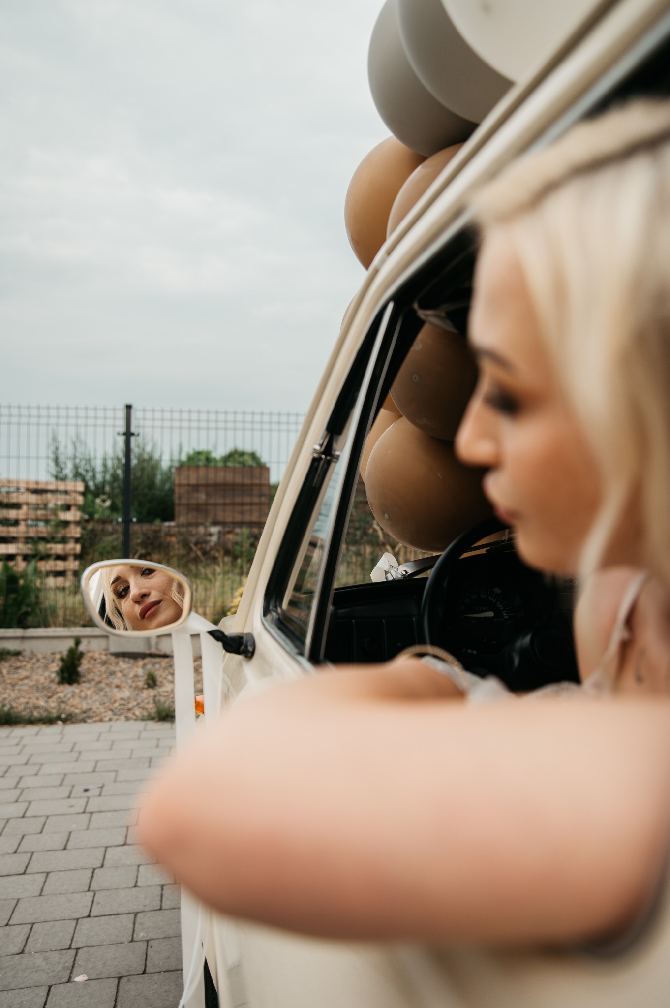 Panna młoda w samochodzie retro ozdobionym beżowymi balonami, jej odbicie w lusterku bocznym, delikatny makijaż i fryzura z warkoczykami.