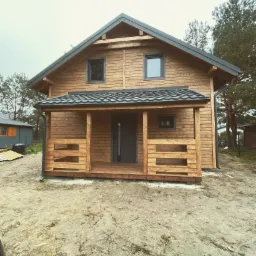 Nowoczesny, parterowy dom z drewna, zadaszone wejście z balustradą, szara stolarka okienna i dachówka, widok z zewnątrz w pochmurny dzień.