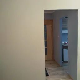 Świeżo pomalowane wnętrze z widokiem na kuchnię przez przejście w ścianie, ściany w odcieniach szarości i beżu, drewniana podłoga i drzwi.