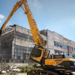 Żółta koparka Hyundai 520LC-9S podczas rozbiórki szarego, betonowego budynku z licznymi oknami, z widocznym pyłem i gruzem w powietrzu.