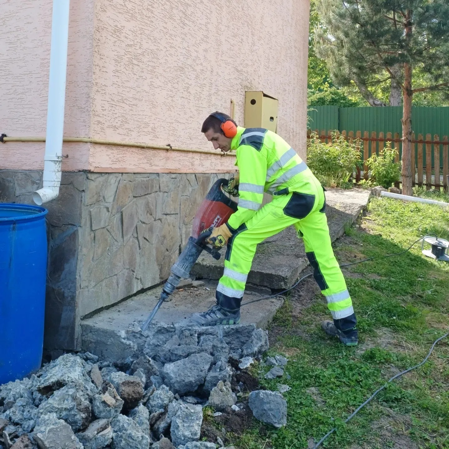 Pracownik w neonowo-zielonym kombinezonie i pomarańczowych słuchawkach ochronnych używa młota pneumatycznego do rozbijania betonowego fundamentu przy budynku z kamienną podmurówką, obok leżą...