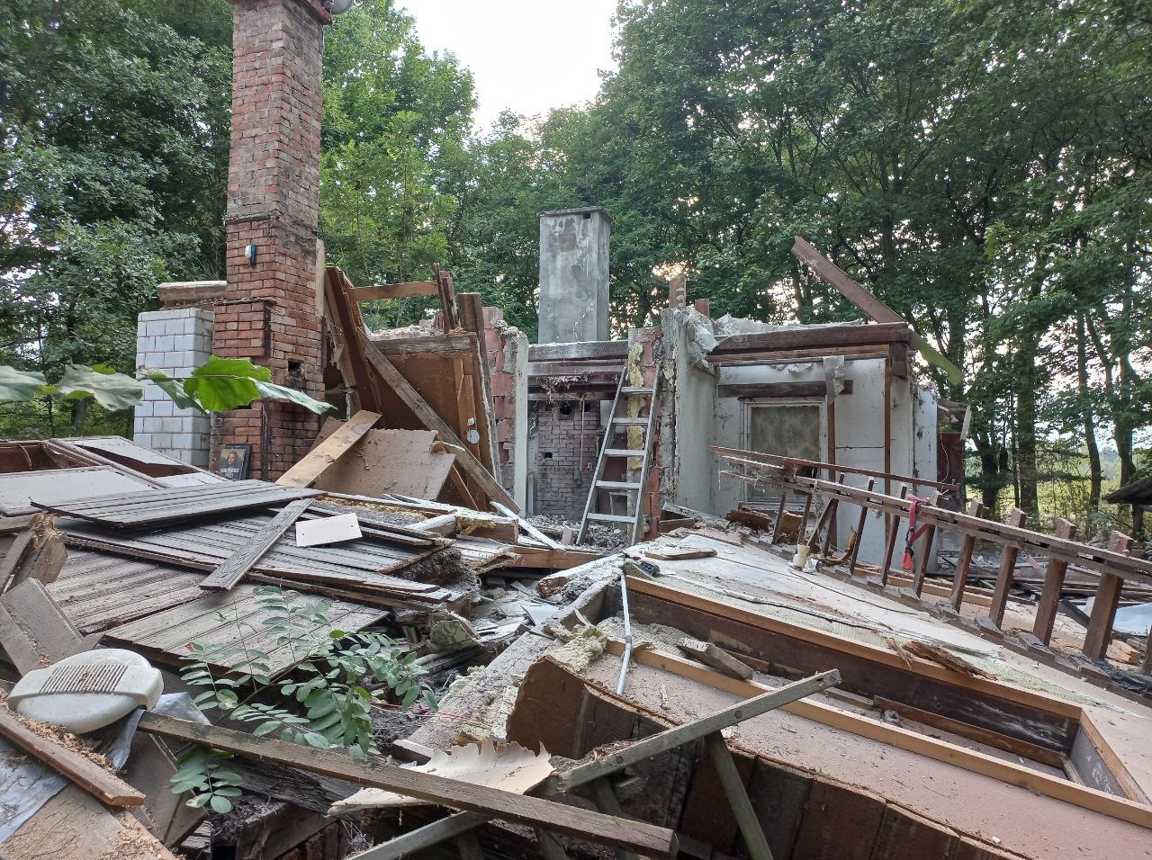 Częściowo zburzony budynek z ceglanym kominem i fragmentami ścian, otoczony zielenią, z widoczną drabiną opartą o ścianę.