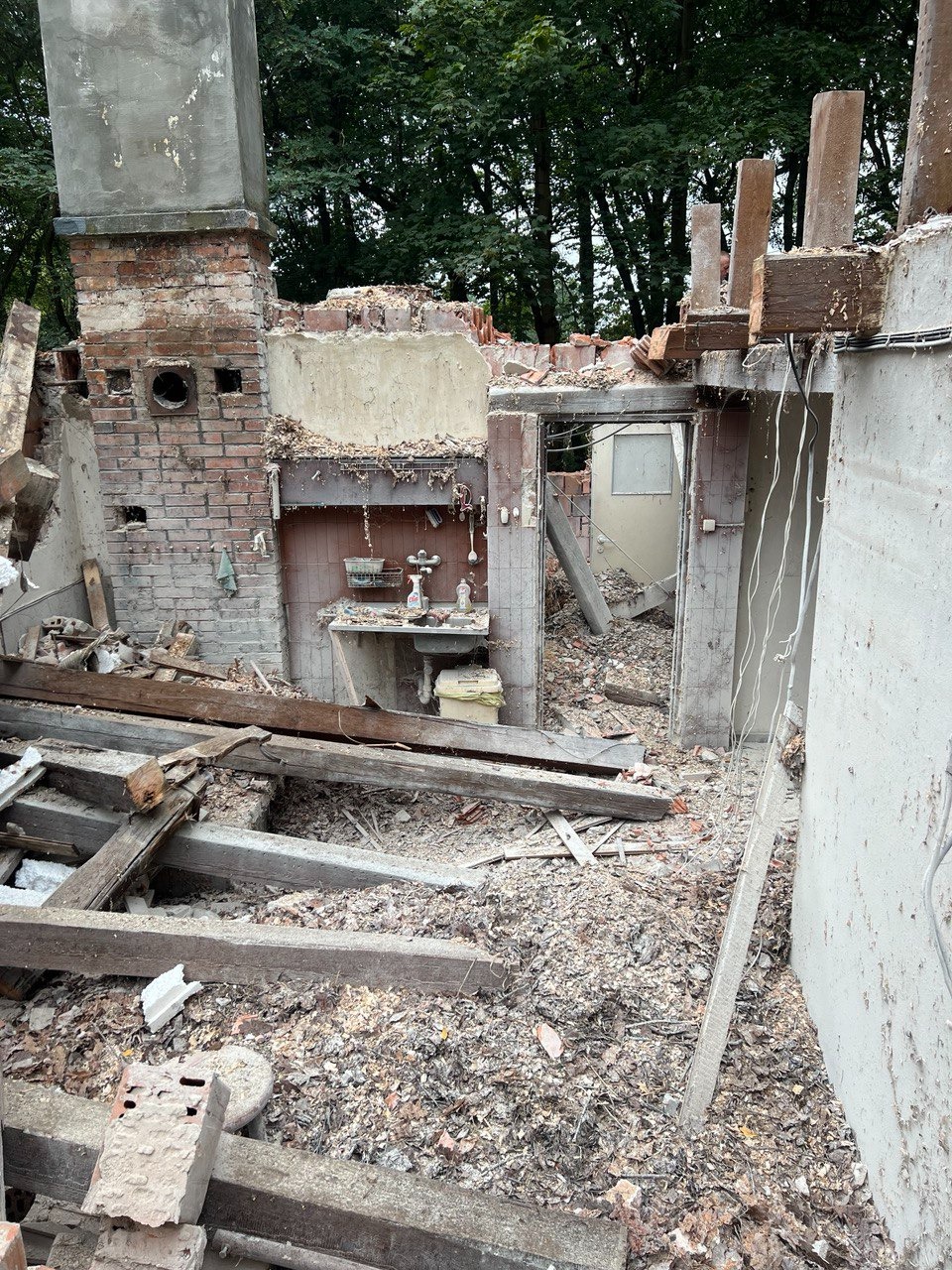 Wnętrze zrujnowanego budynku po rozbiórce, widoczna pozostałość ceglanego komina, fragmenty ścian, gruz i drewniane belki na podłodze, zieleń drzew w tle.