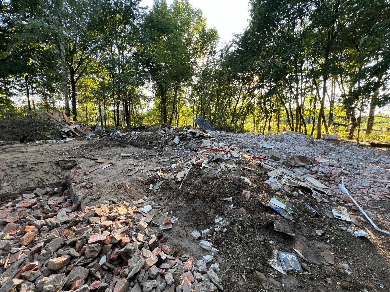 Teren po rozbiórce budynku z widocznymi gruzami ceglanymi, fragmentami drewna i odpadami budowlanymi na tle drzew w słoneczny dzień.