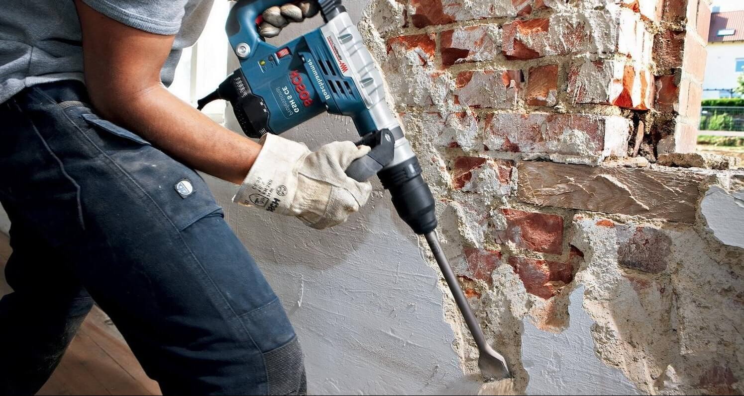 Pracownik w rękawicach roboczych używa młota udarowego Bosch do skuwania tynku ze ściany z czerwonej cegły podczas prac remontowych.