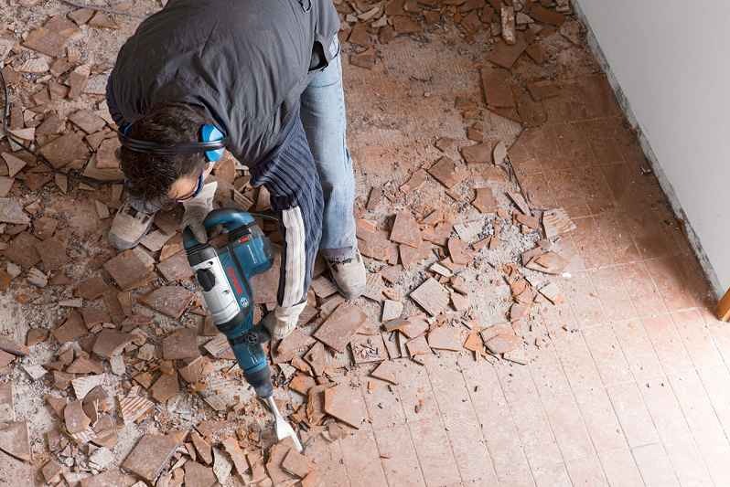 Pracownik w słuchawkach ochronnych skuwa starą, brązową podłogę z płytek za pomocą młota udarowego Bosch, widok z góry na gruz i fragment ściany.