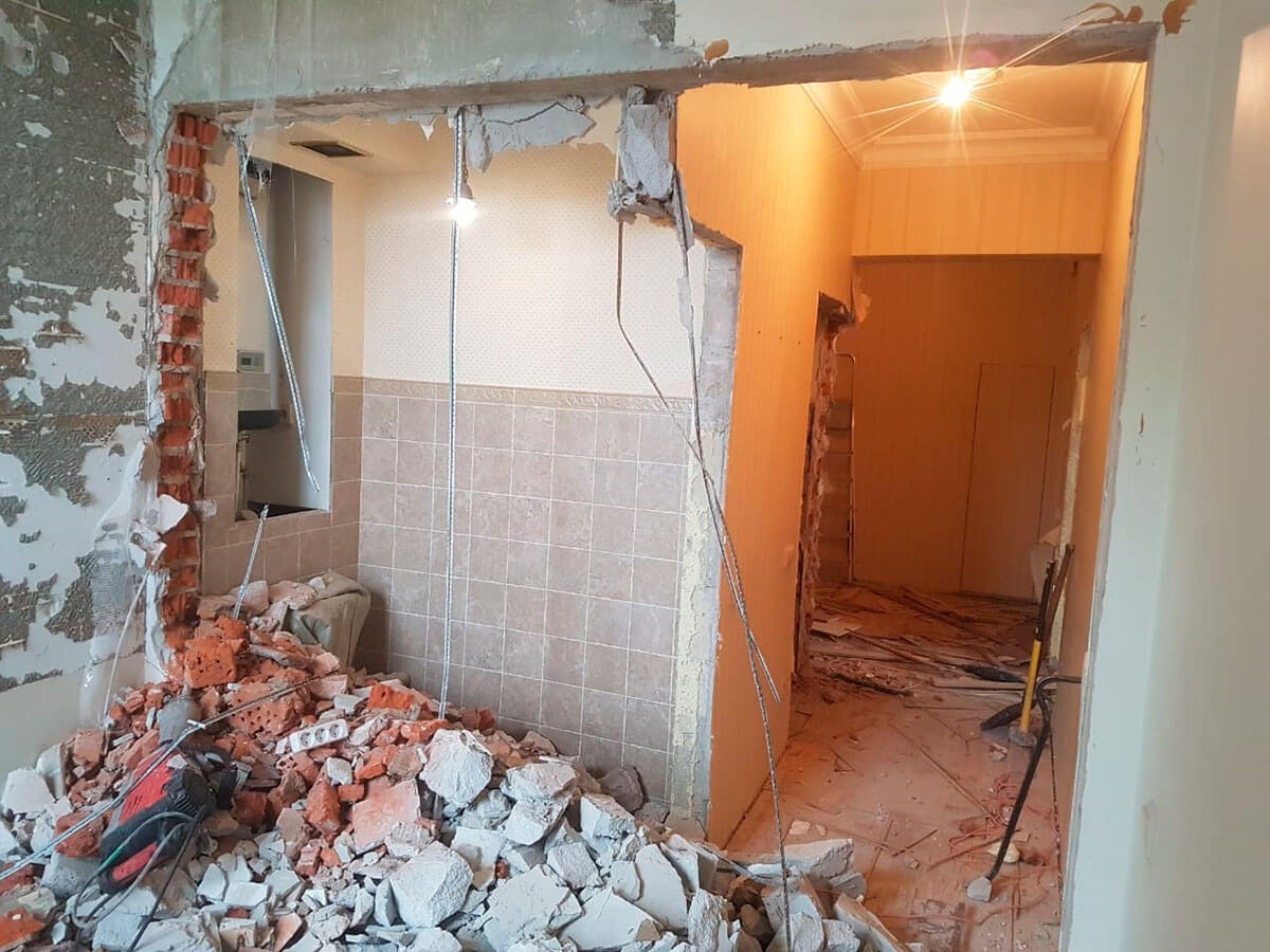 Częściowo wyburzona ściana działowa z widocznymi cegłami i gruzem, narzędzie do wyburzania leżące obok, widok na sąsiednie pomieszczenie z resztkami gruzu na podłodze.