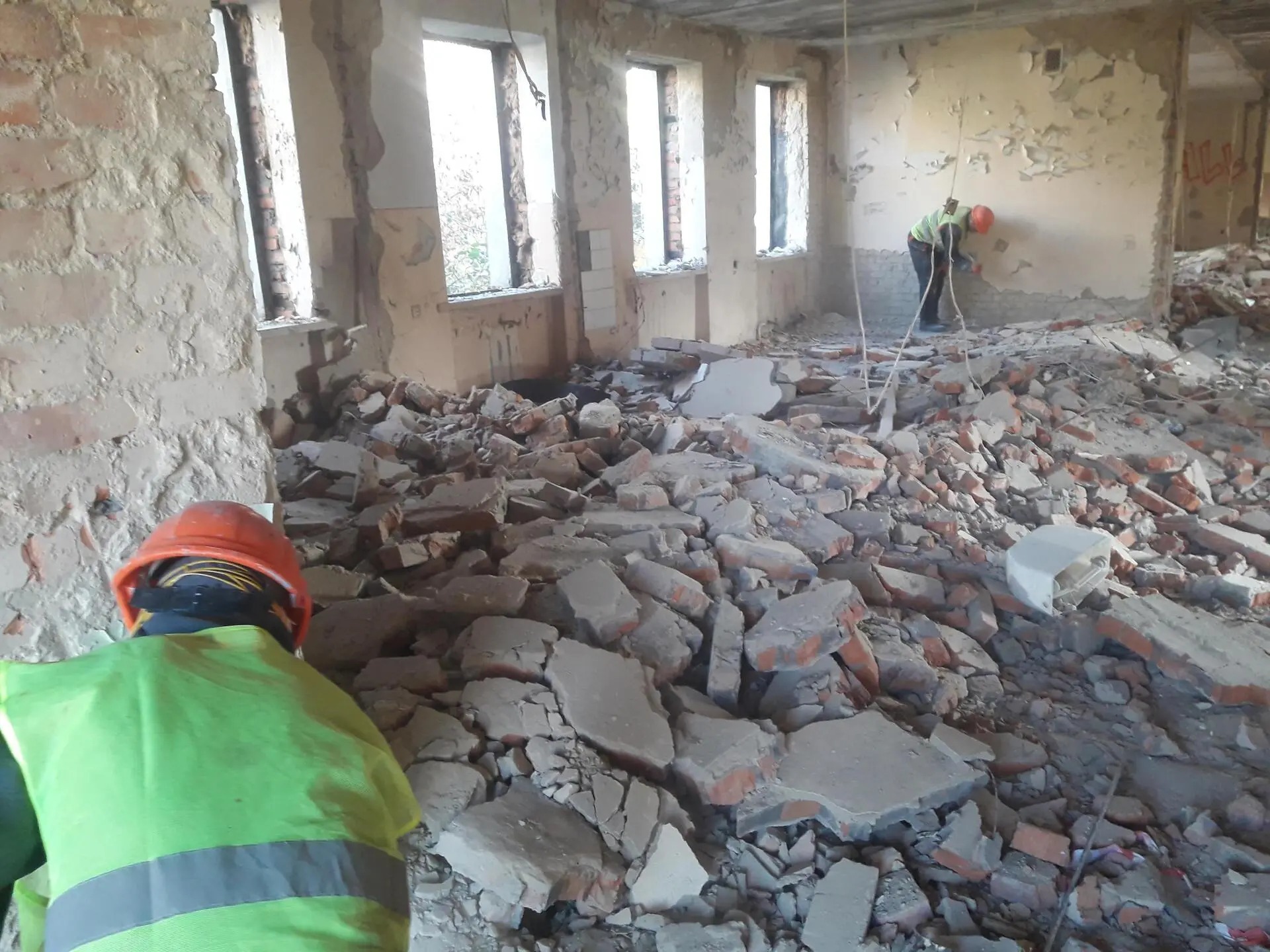 Wnętrze budynku w trakcie rozbiórki; gruzowisko z cegieł i betonu, pracownik w kasku i kamizelce odblaskowej usuwa resztki tynku ze ściany, widoczne okna bez szyb.