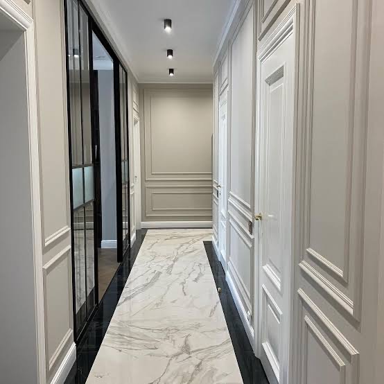 Elegancki korytarz w odcieniach szarości i bieli z marmurową podłogą, czarnymi ramami szklanych drzwi i ozdobnymi panelami ściennymi. Nowoczesne oświetlenie punktowe.