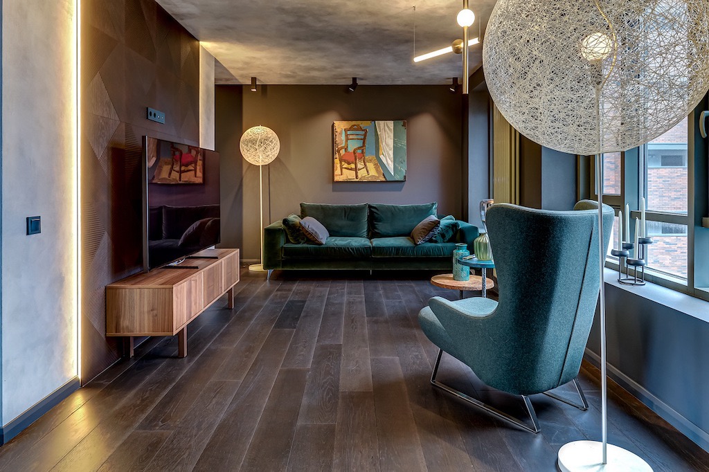 Stylowy salon z ciemną podłogą, zieloną sofą i fotelem. Dekoracyjna ściana z podświetleniem LED. Obraz nad sofą. Nowoczesne lampy i świece przy oknie.