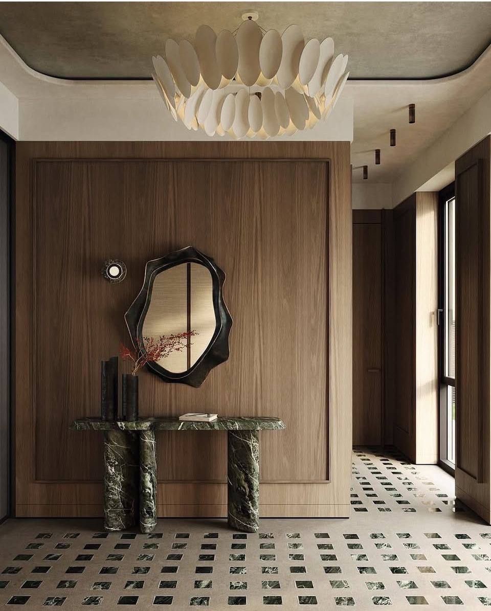Elegancki hol z drewnianą boazerią, marmurową konsolą i lustrem o nieregularnym kształcie. Dekoracyjna lampa sufitowa i wzorzysta podłoga dodają wnętrzu charakteru.