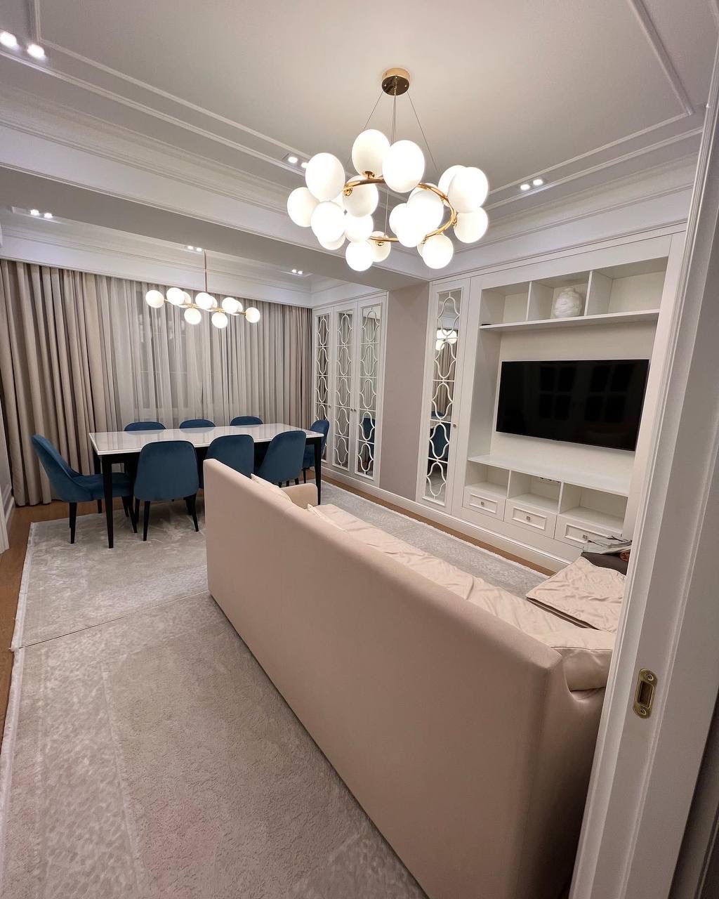 Elegancki salon z beżową sofą, niebieskimi krzesłami przy stole i nowoczesnym oświetleniem. Białe meble w zabudowie i jasny dywan dopełniają aranżację.