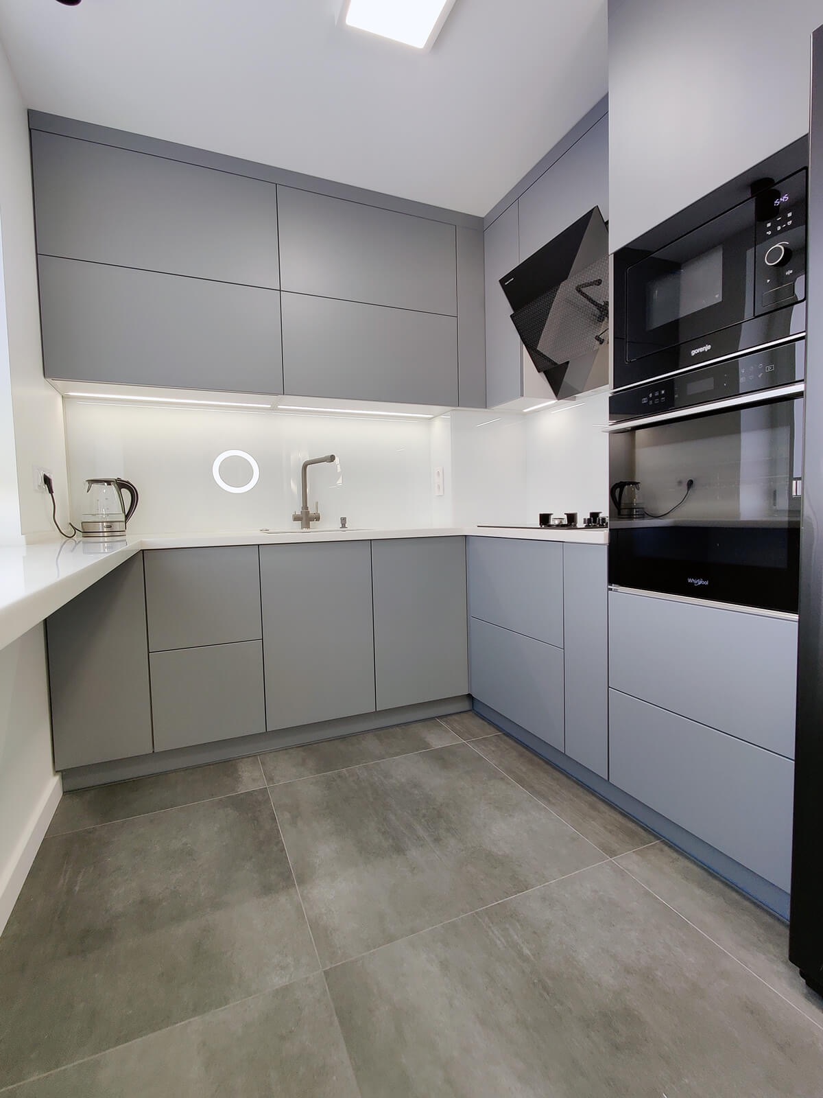 Nowoczesna, szara kuchnia z oświetleniem LED pod szafkami, płytkami podłogowymi w odcieniach szarości i zabudowanym sprzętem AGD.