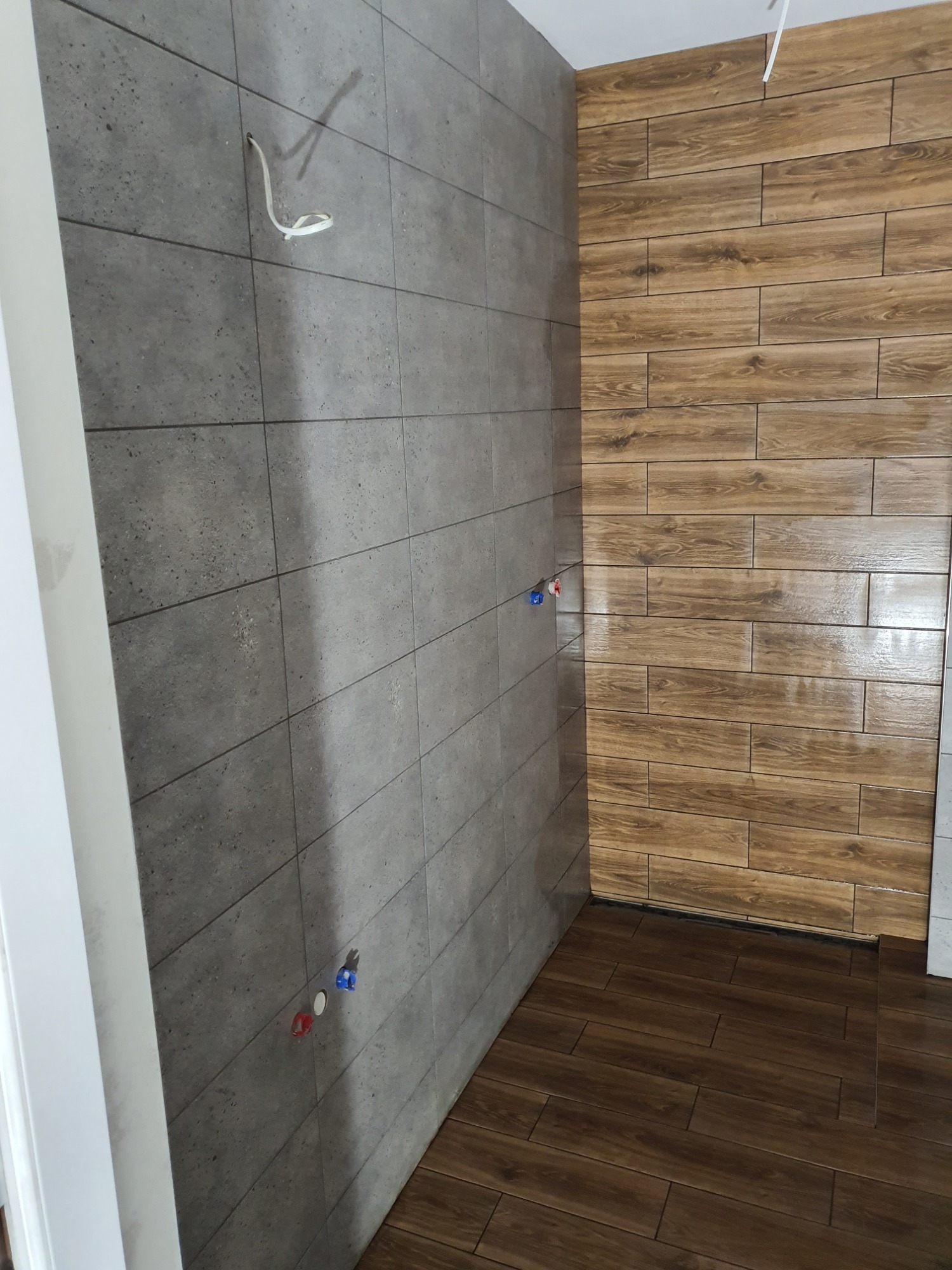 Wnętrze łazienki w trakcie wykańczania: szare, matowe płytki ścienne z widocznymi przyłączami wodnymi i wystającym kablem, oraz drewnopodobne płytki na ścianie i podłodze ułożone w poziome pasy.