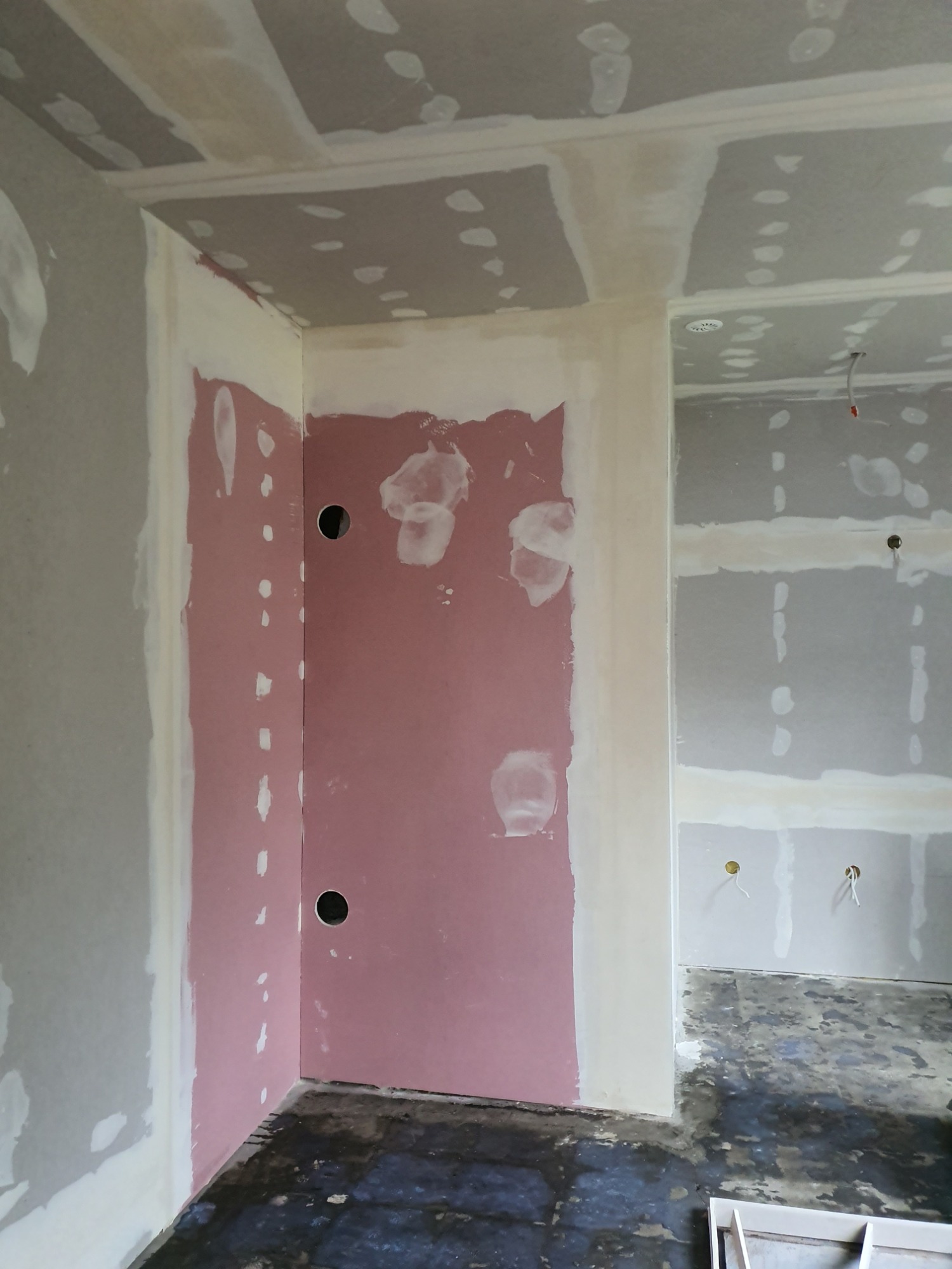 Wnętrze w trakcie remontu: ściany z płyt gipsowo-kartonowych, częściowo zagruntowane, widoczne otwory na instalacje elektryczne, surowa podłoga.