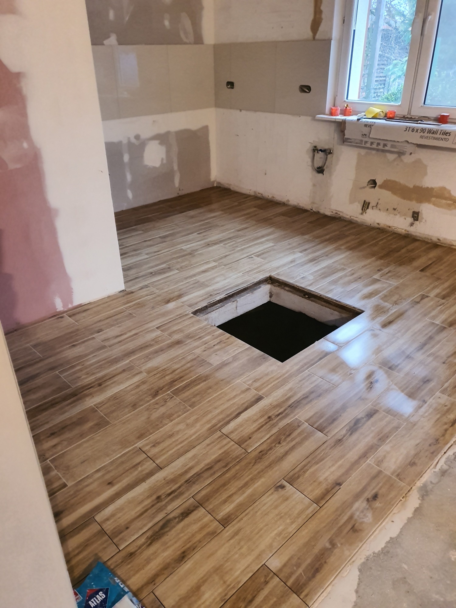 Układanie podłogi z płytek imitujących drewno w trakcie remontu, widoczny otwór w podłodze, ściany przygotowane do malowania, karton z płytkami na parapecie.