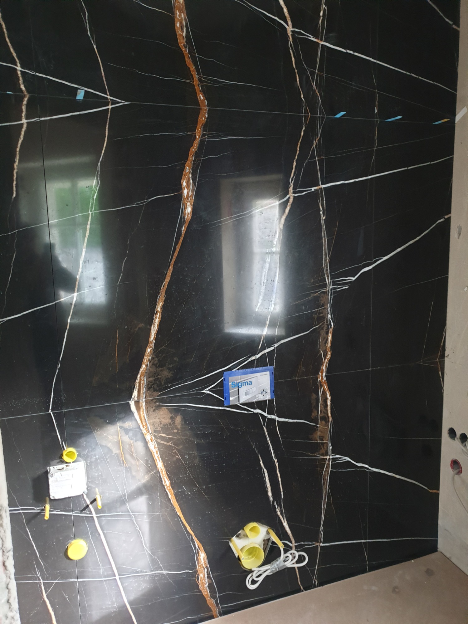 Ściana wyłożona czarnymi płytkami imitującymi marmur z białymi i złotymi żyłami, widoczne elementy instalacji hydraulicznej w trakcie montażu.