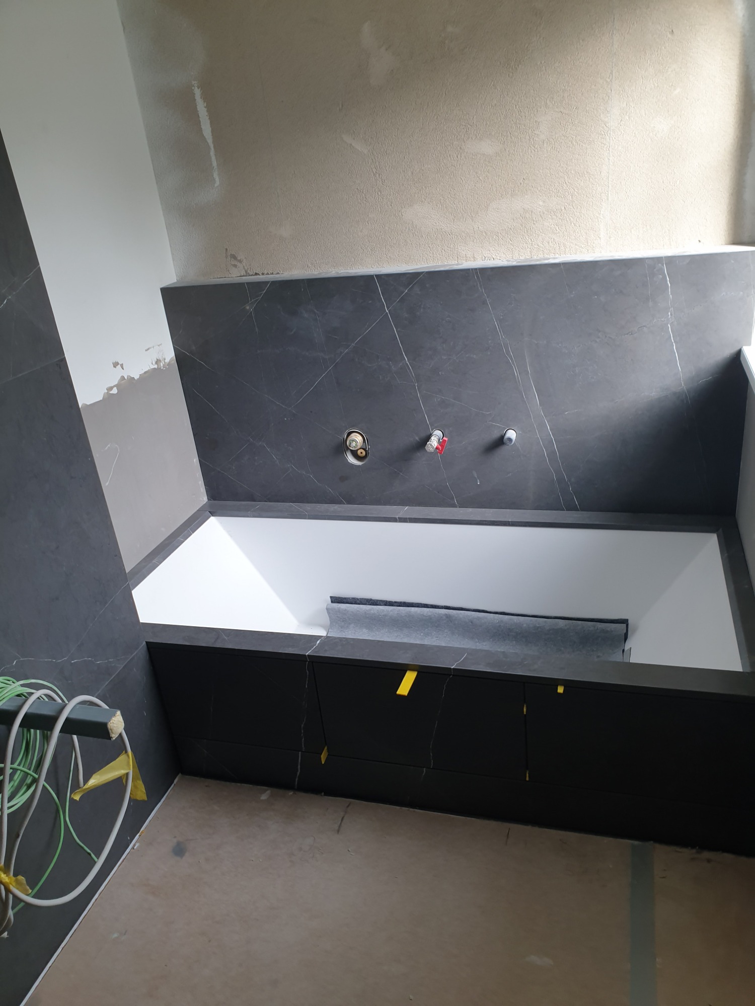 Nowoczesna biała wanna obudowana czarnymi płytkami imitującymi marmur, widoczne przygotowane instalacje wodne i fragment nie wykończonej ściany, sugerujące etap prac wykończeniowych.