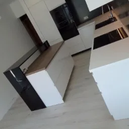 Nowoczesna, biała kuchnia z wyspą, drewnianym blatem i czarnymi akcentami, widok z niskiego kąta, jasna podłoga.