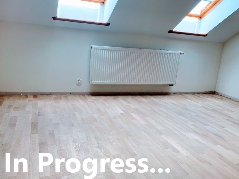 Jasne, drewniane deski podłogowe ułożone w jodełkę, widoczne ślady po listwach przypodłogowych, grzejnik ścienny i okna dachowe w jasnym pokoju, etap w trakcie realizacji.