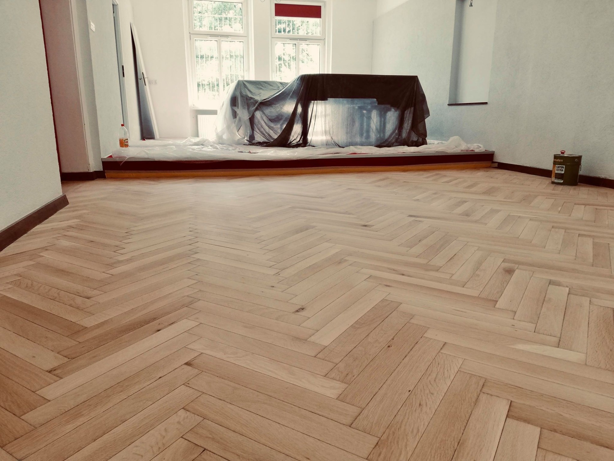 Jasny parkiet ułożony we wzór jodełki francuskiej w pokoju z oknami, zabezpieczony fortepian pod folią ochronną i puszka lakieru przy ścianie.