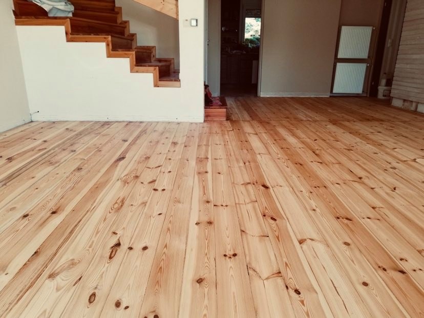 Świeżo wycyklinowana sosnowa podłoga w jasnym odcieniu z widocznymi słojami, sąsiadująca ze schodami z drewnianymi stopniami, w tle grzejnik.