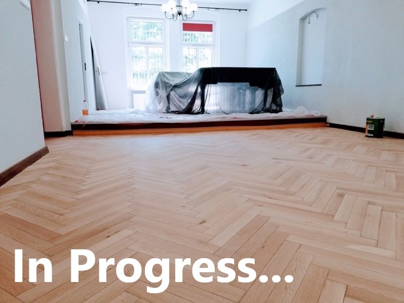 Jasny parkiet ułożony w jodełkę w trakcie renowacji, zabezpieczone meble pod czarną folią, puszka farby przy ścianie.