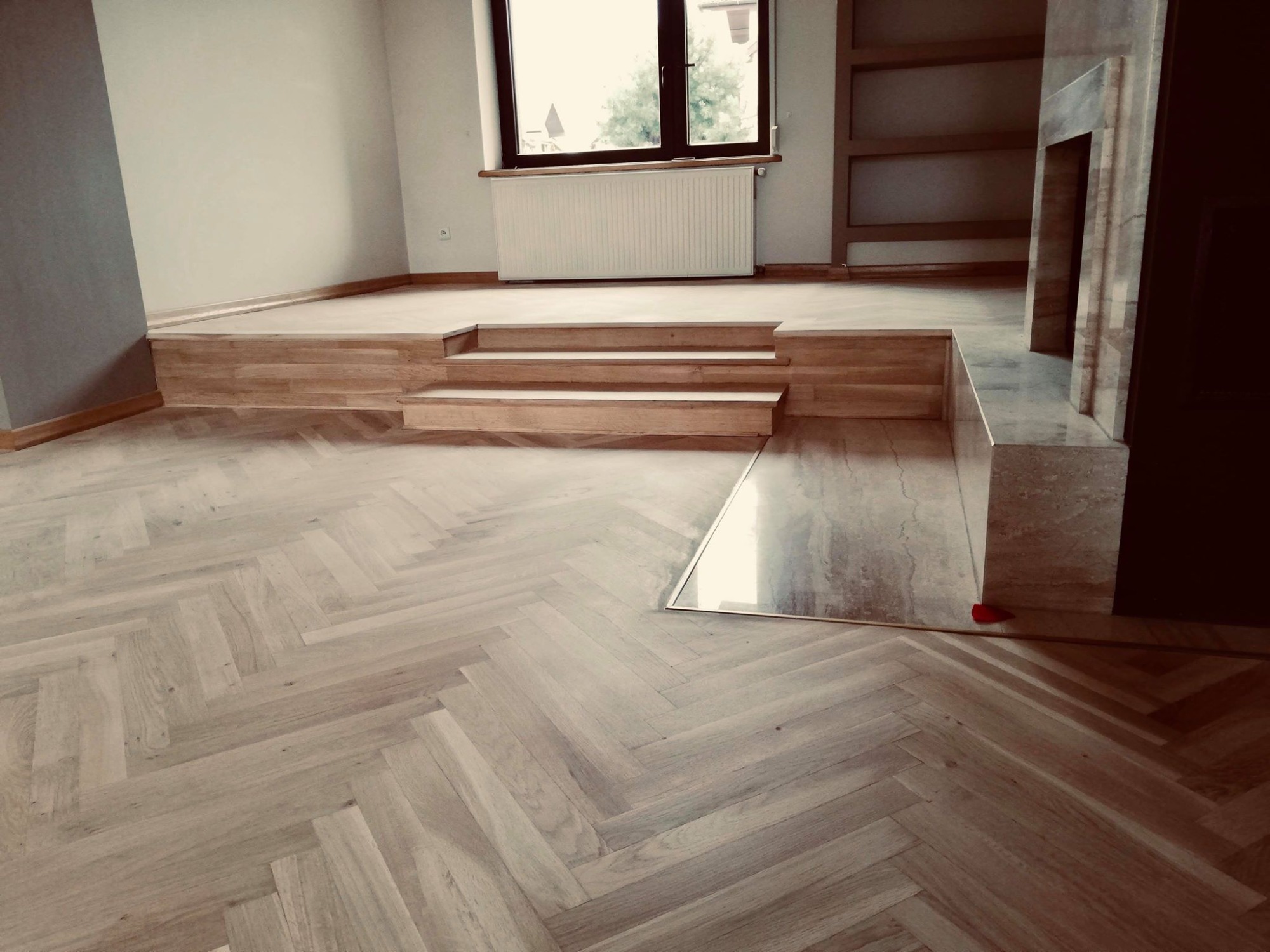 Jasny parkiet ułożony we wzór jodełki francuskiej, z drewnianymi schodkami prowadzącymi do podwyższonej części podłogi i marmurowym obramowaniem kominka.