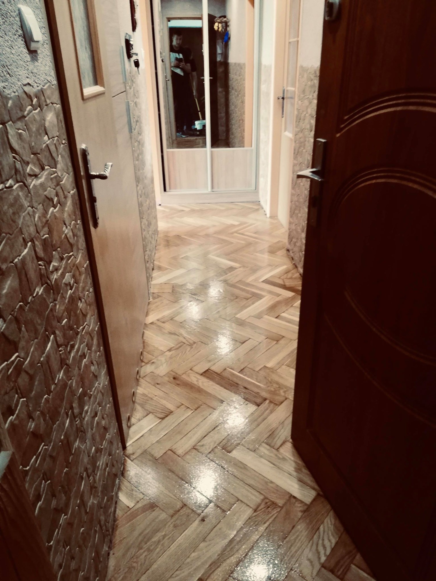 Korytarz z parkietem ułożonym w jodełkę, widoczne drzwi po lewej stronie z dekoracyjną fakturą na ścianie oraz otwarte drzwi po prawej stronie, w tle widoczna postać z mopem.