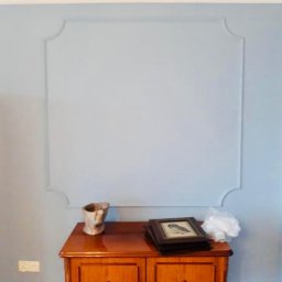 Renow Obrebski - Ściana pomalowana na jasny niebieski kolor z ozdobnym kwadratem z listew, poniżej drewniana komoda z dekoracjami.