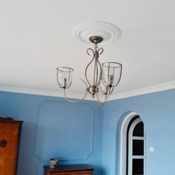 Renow Obrebski - Pomalowane na niebiesko ściany pokoju z dekoracyjną sztukaterią, klasyczna szafka i komoda z ciemnego drewna, oraz żyrandol z transparentnymi kloszami